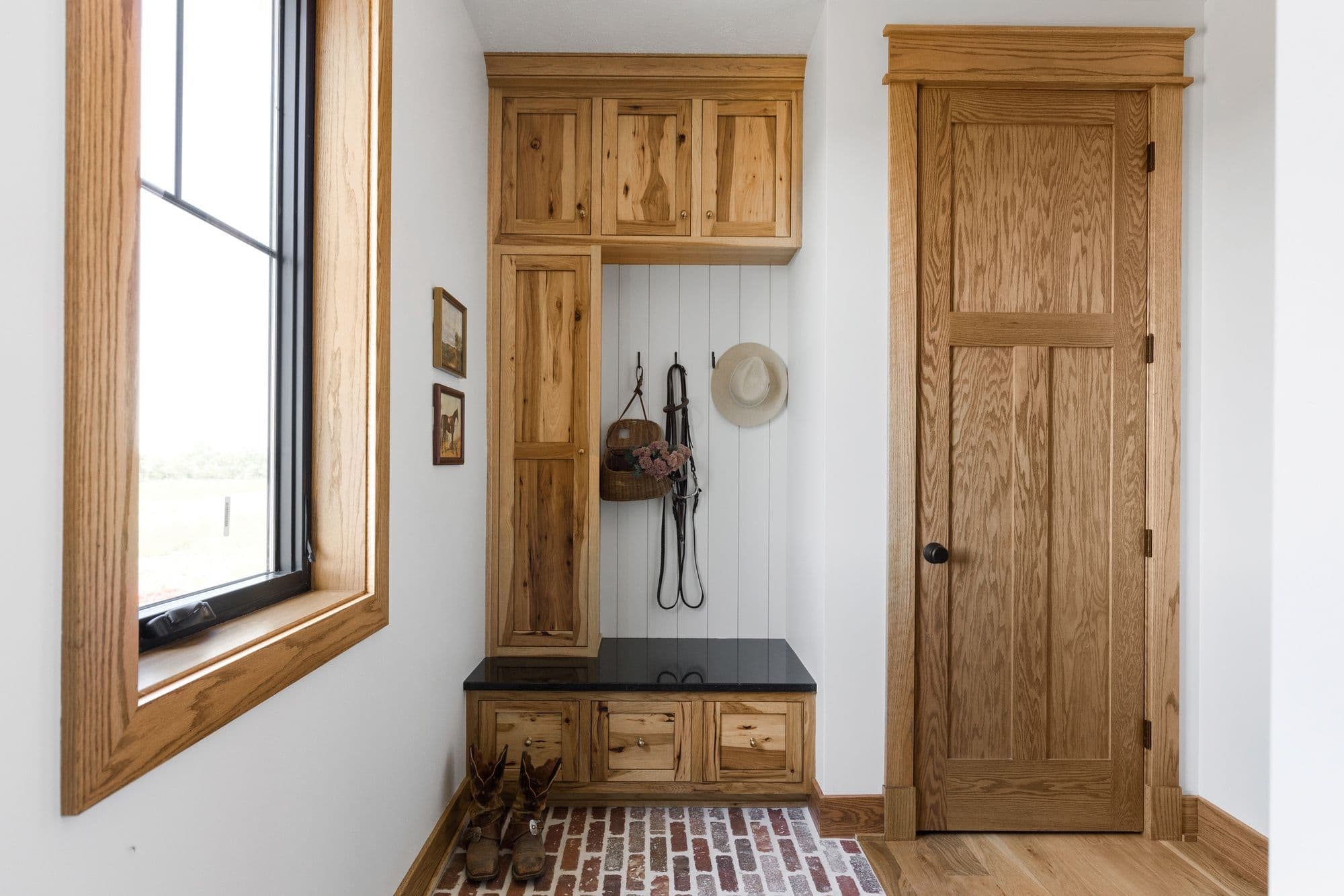 Entry mudroom with natural wood cabinetry featuring open shelving, storage drawers, and a black countertop bench. A window with wood trim sits to the left, and a paneled wood door to the right, all on a brick floor.