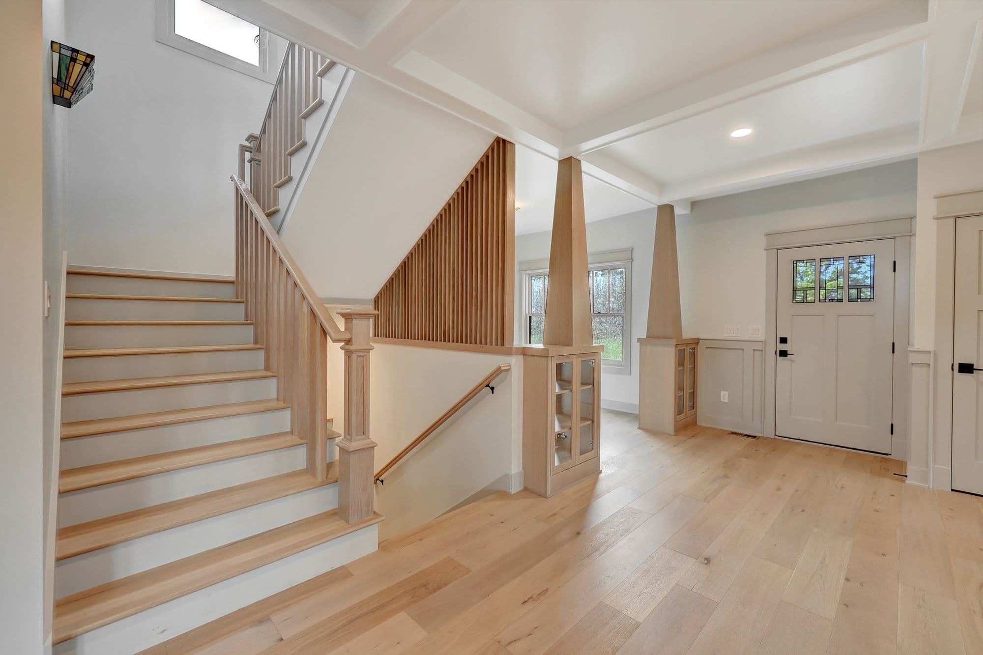 Interior entryway featuring hardwood floors, a staircase with wooden railings, and a front door with decorative glass panels. The space is well-lit with natural light filtering through windows.