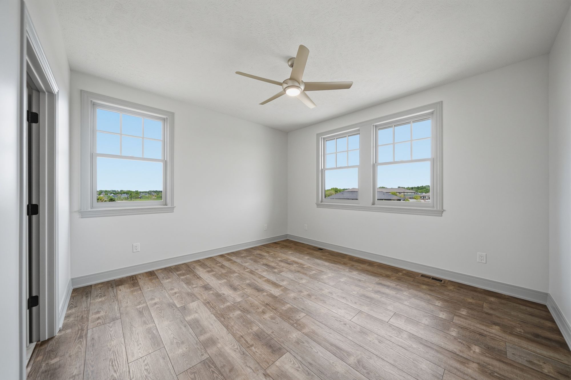 Bright bedroom featuring hardwood floors, two windows providing natural light, and a ceiling fan. Walls are painted a soft white and the windows are trimmed in white molding.