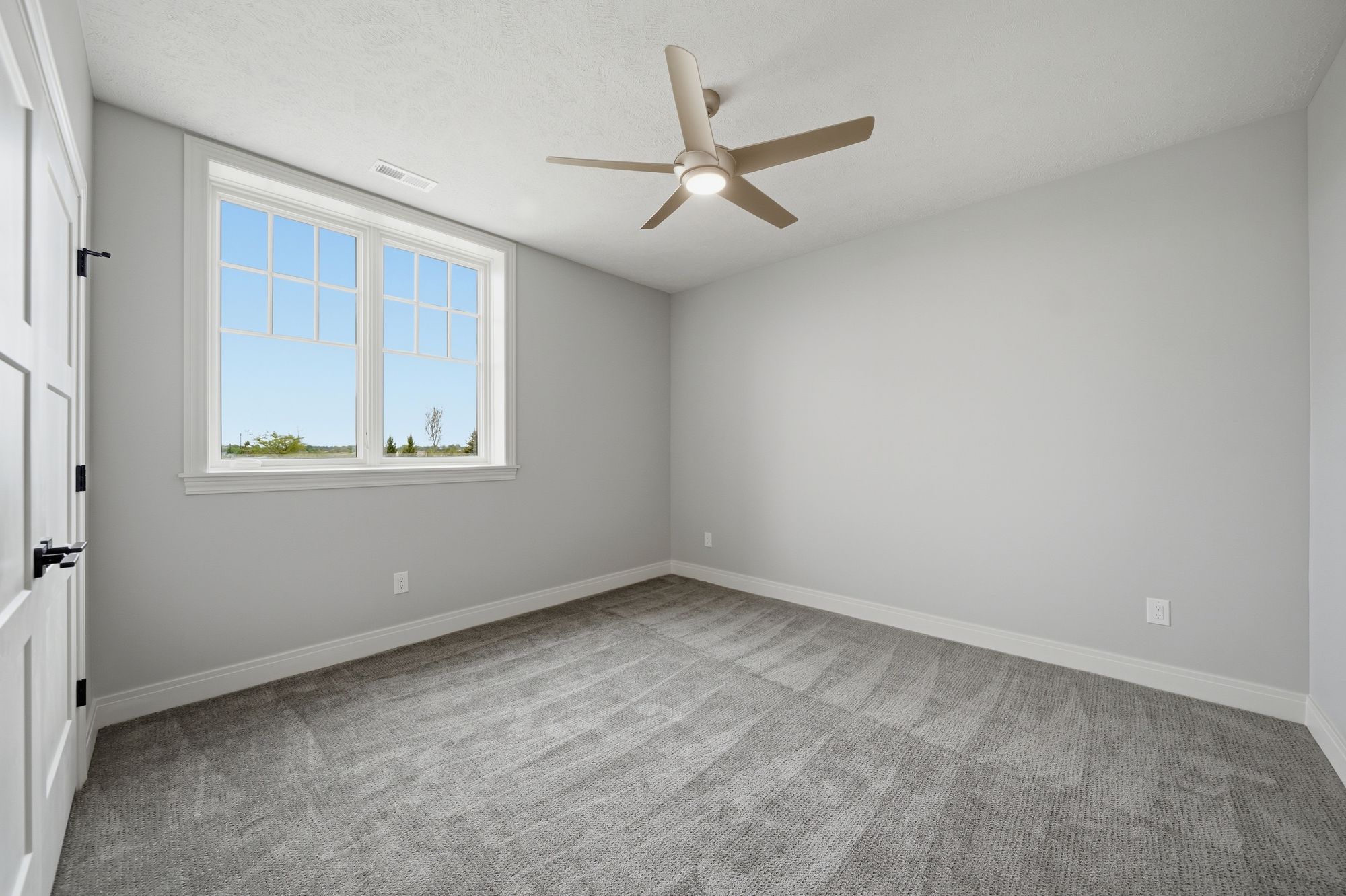 A bedroom features gray carpet and walls, a white-framed window, and a beige ceiling fan with a light fixture. The room is mostly empty, providing an open space.