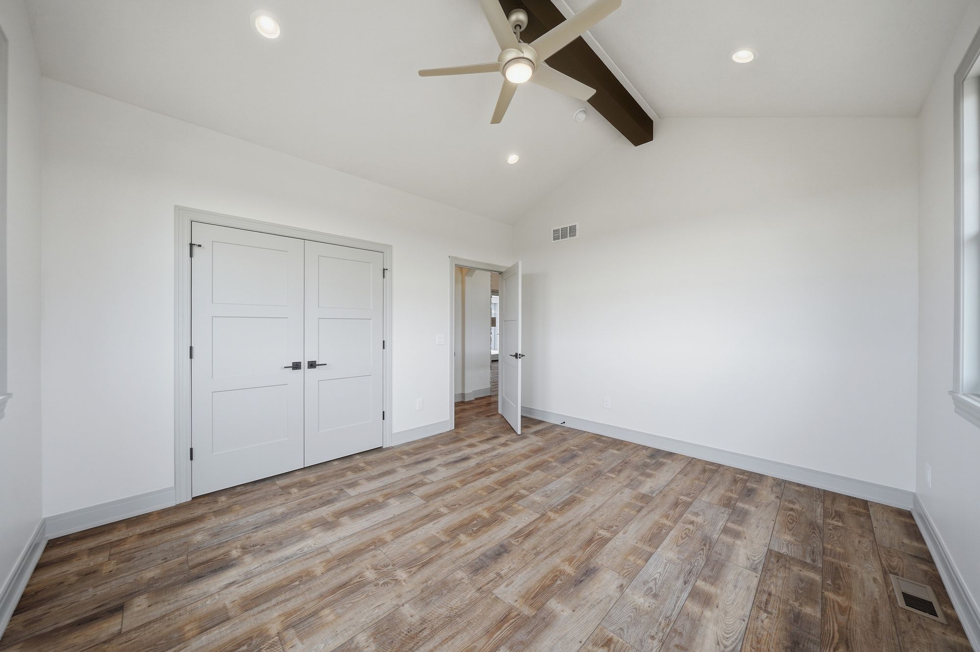 Bright bedroom features light wood-look flooring, white walls, and double closet with square panel doors. The room has a ceiling fan, recessed lighting, and a brown ceiling beam.