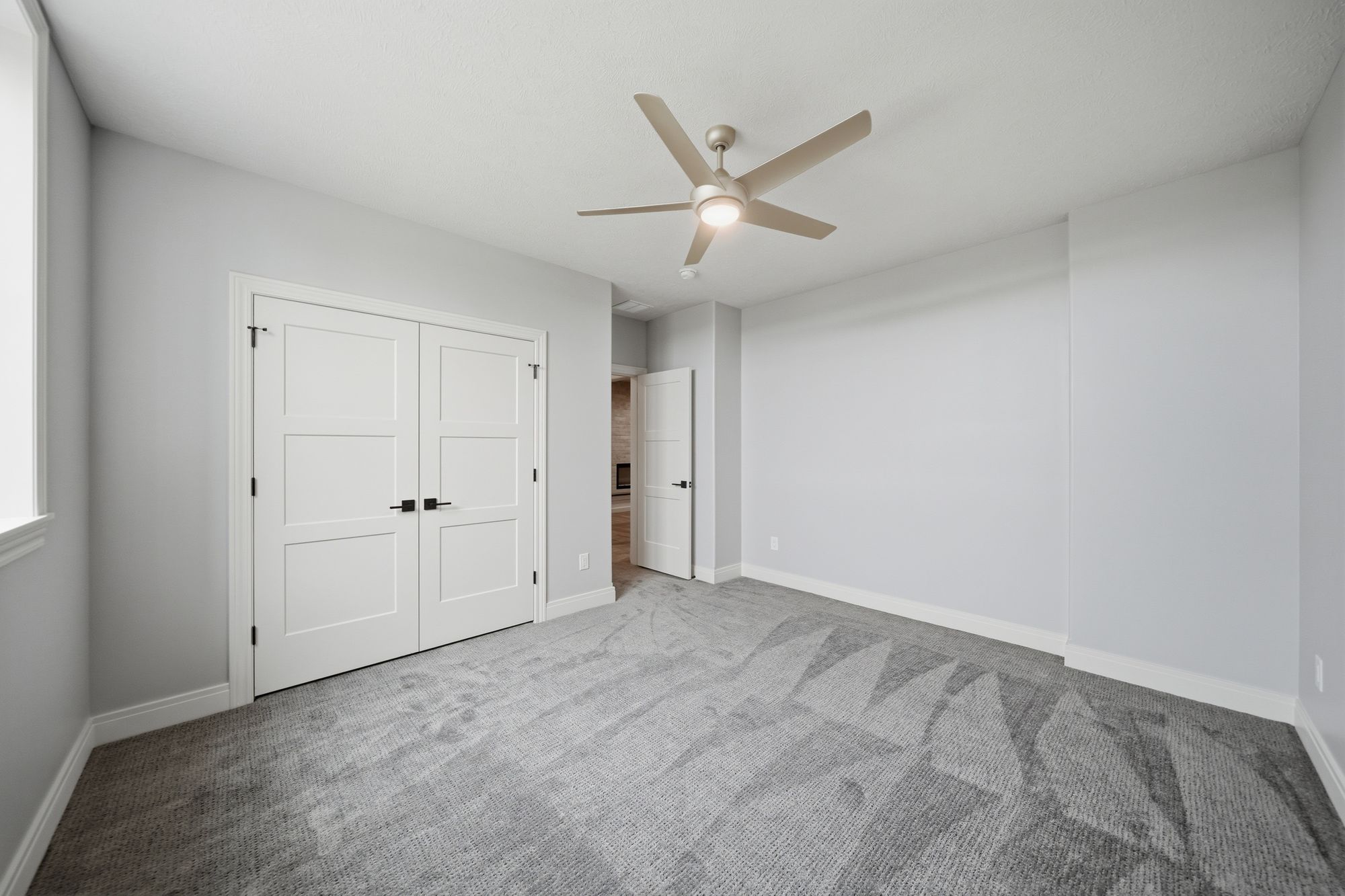 Empty bedroom featuring gray carpet, light gray walls, and white trim. A ceiling fan is centered in the room, which also has a closet with paneled white doors and black hardware.