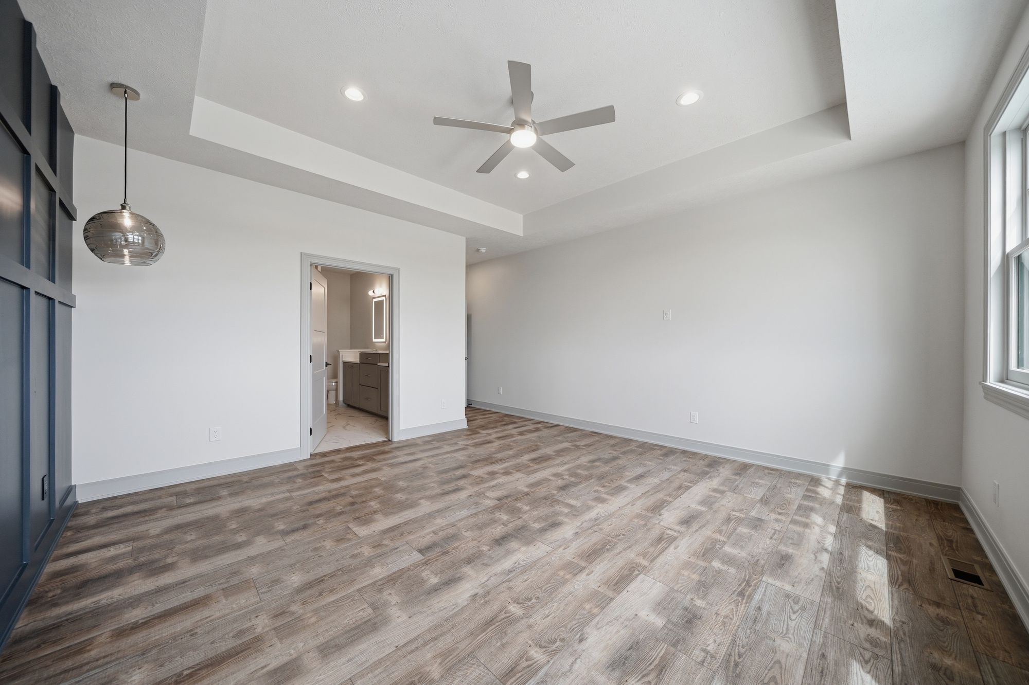Spacious bedroom with gray-toned wood floors, recessed lighting, and a ceiling fan. The room features a pendant light and an ensuite bathroom visible through an open doorway.