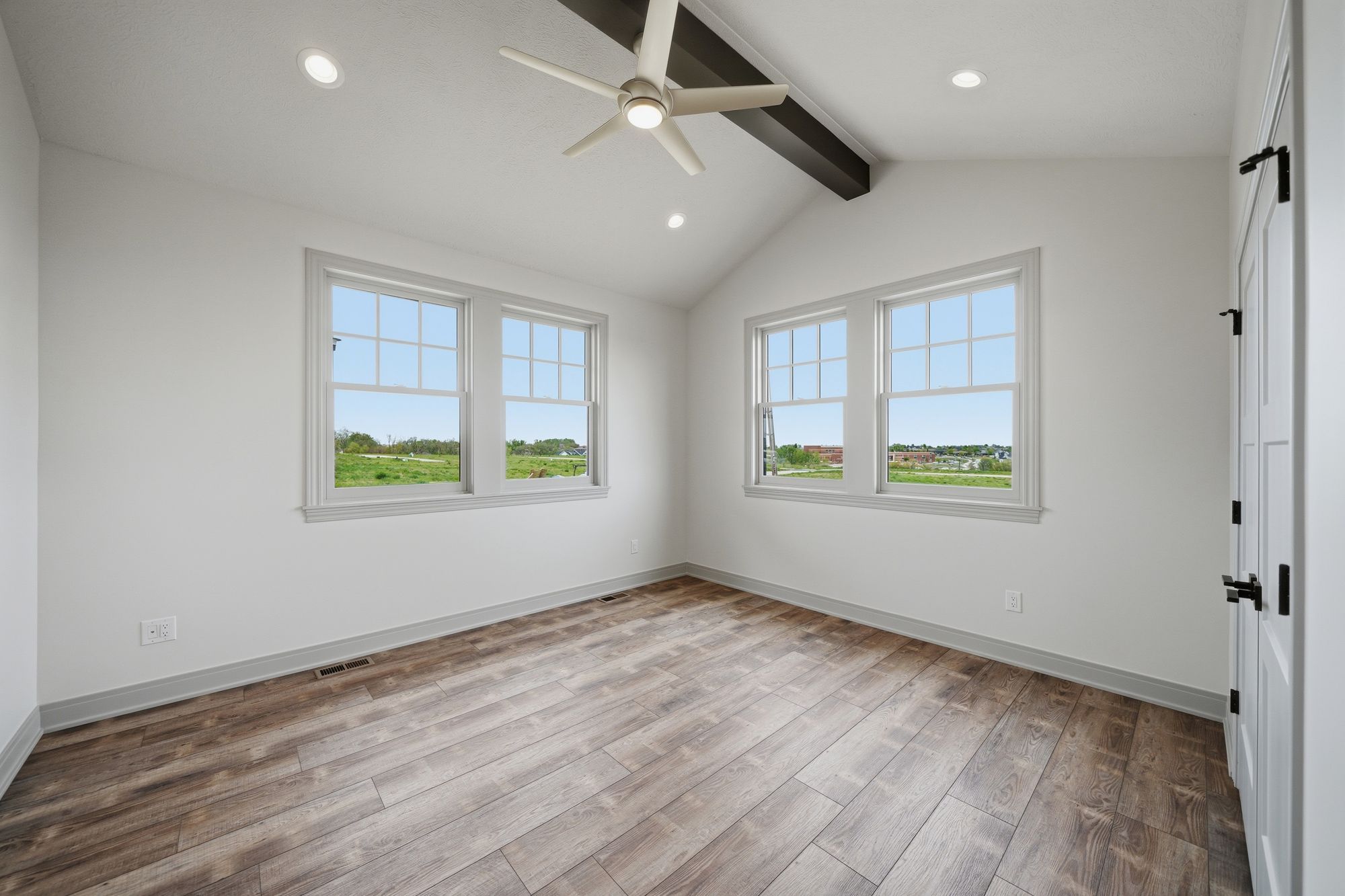 Bedroom featuring two windows with grid patterns offering scenic views, complemented by hardwood floors laid in a diagonal pattern and a white ceiling with a light-colored fan and dark beam accent.