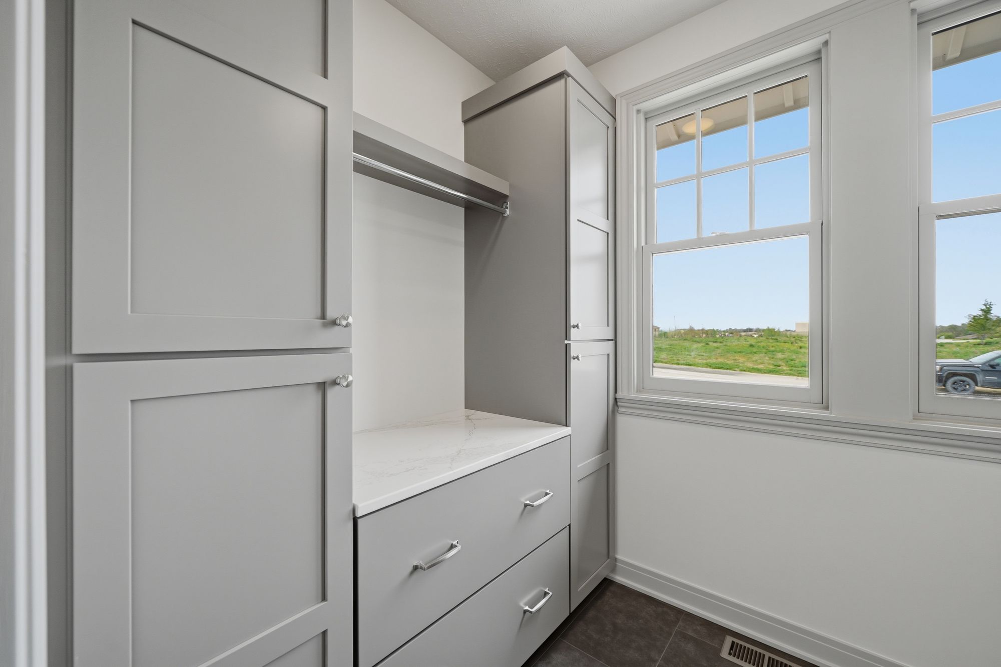This is a closet with gray cabinets, drawers, a clothes bar, and white countertops. A window with a view of a green field and a gray vehicle provides natural light to the space.