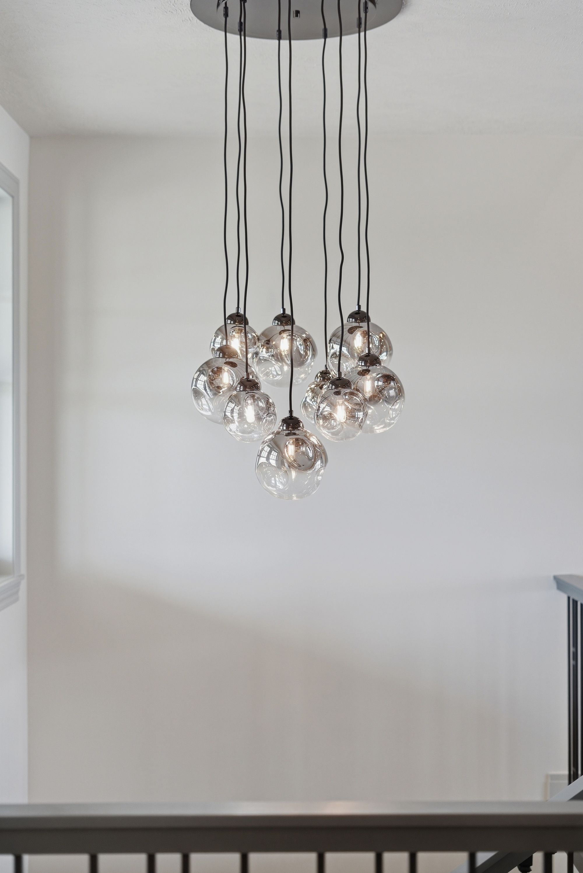 Modern chandelier with multiple clear glass globes hanging from the ceiling. The fixture illuminates a bright hallway or entry space in a home with white walls and a gray railing.