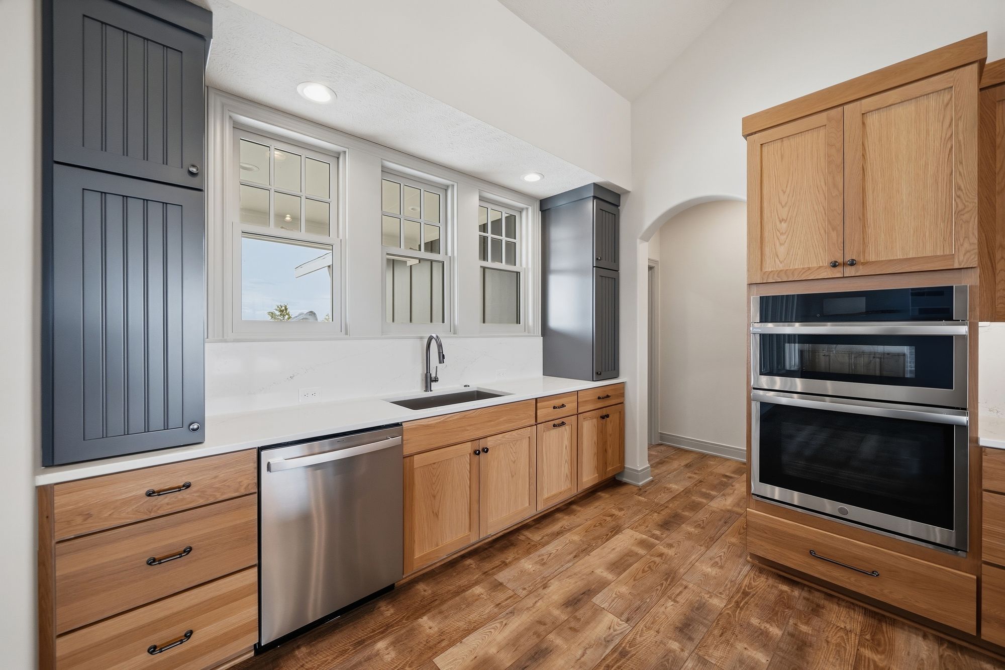 Kitchen featuring hardwood floors, wood cabinets, stainless steel appliances, and a light-colored countertop. There are several windows above the sink, offering natural light.