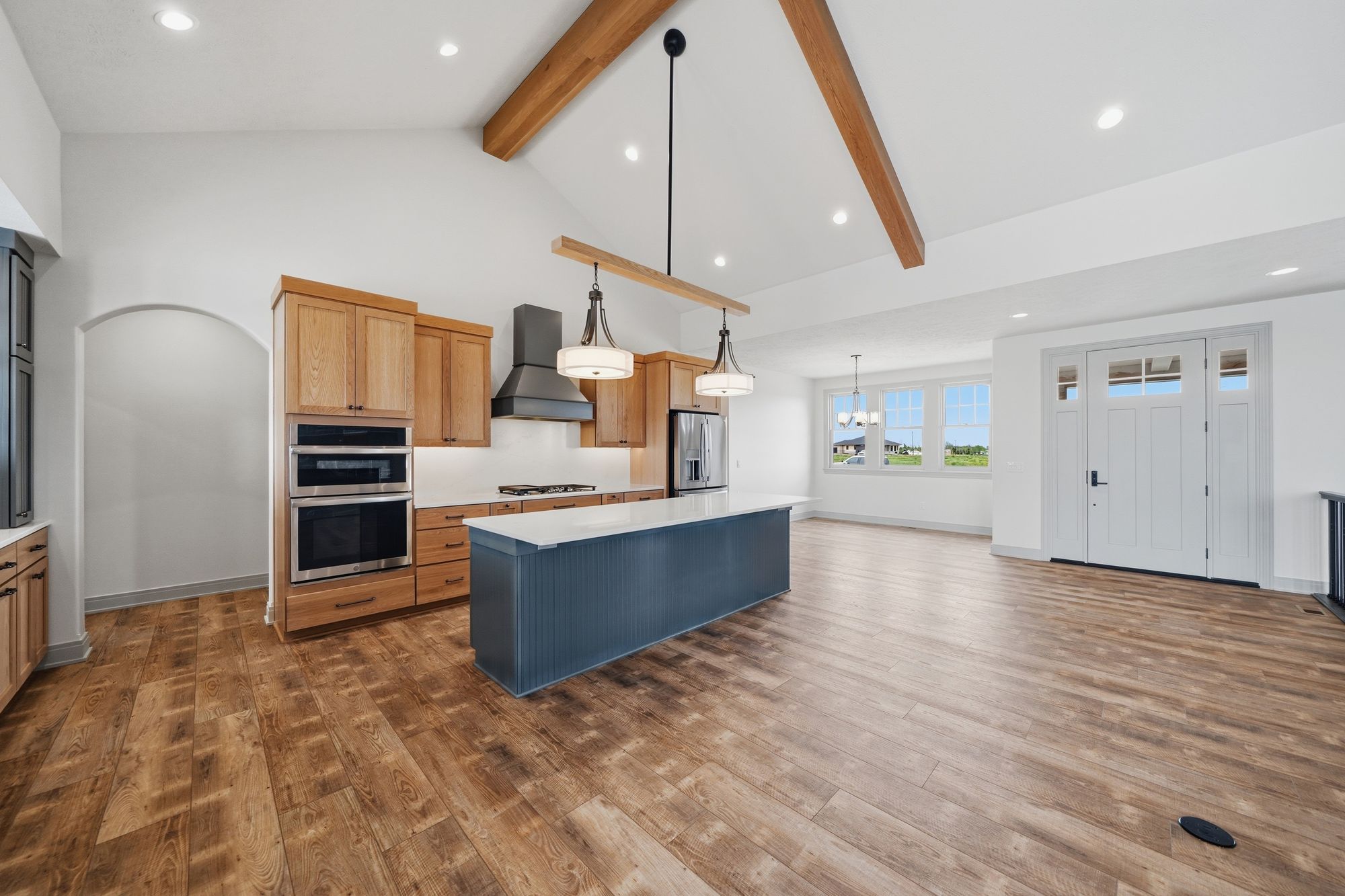 Open concept kitchen features natural wood cabinetry, a navy blue island with a white countertop, and hardwood floors. Wooden beams accent the vaulted ceiling, and stainless steel appliances complement the modern design.