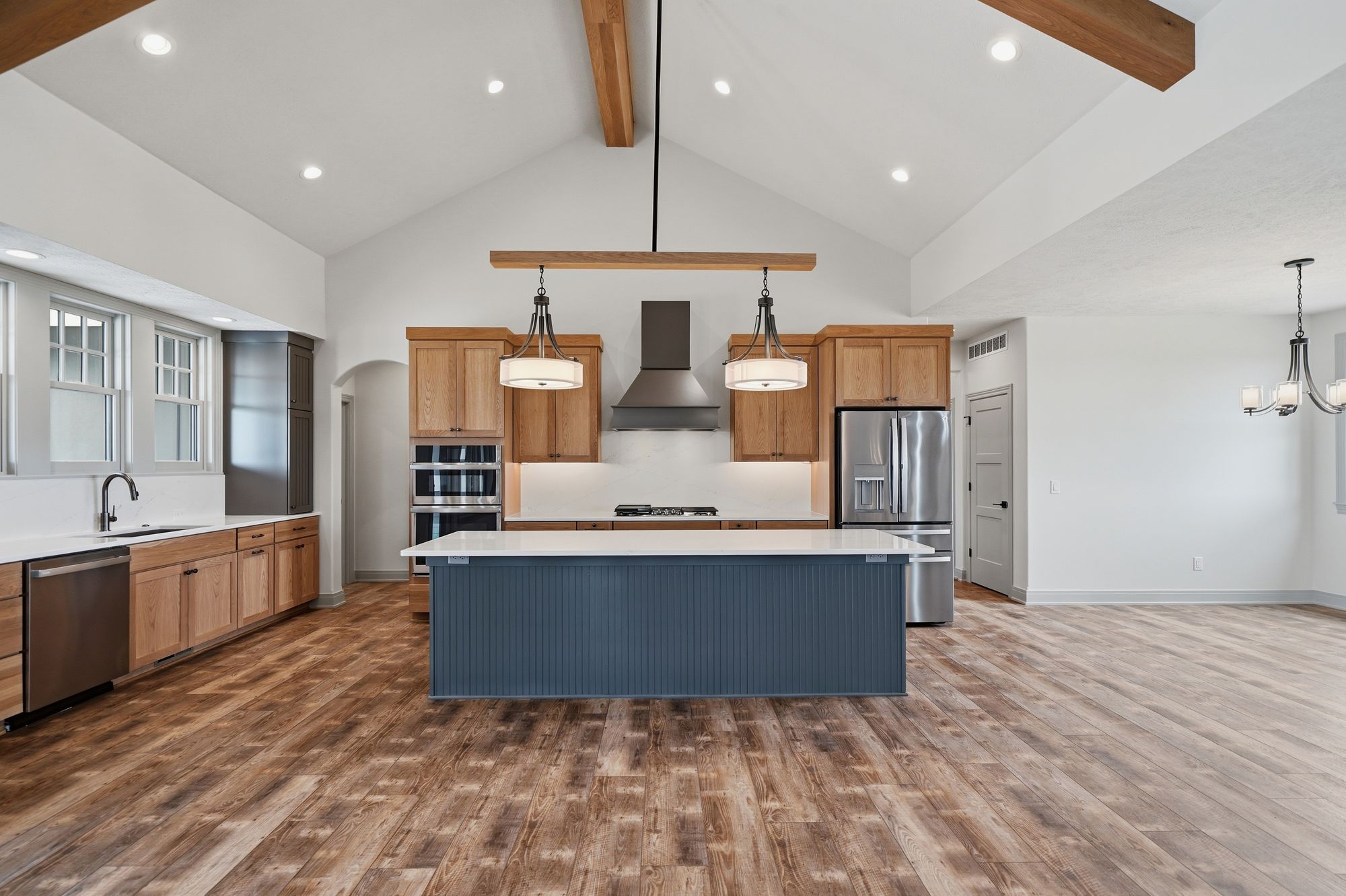 Spacious kitchen featuring a large blue-gray island with white countertop, open floor plan with natural wood floors, wood cabinets, and stainless steel appliances under a vaulted ceiling with wooden beams.