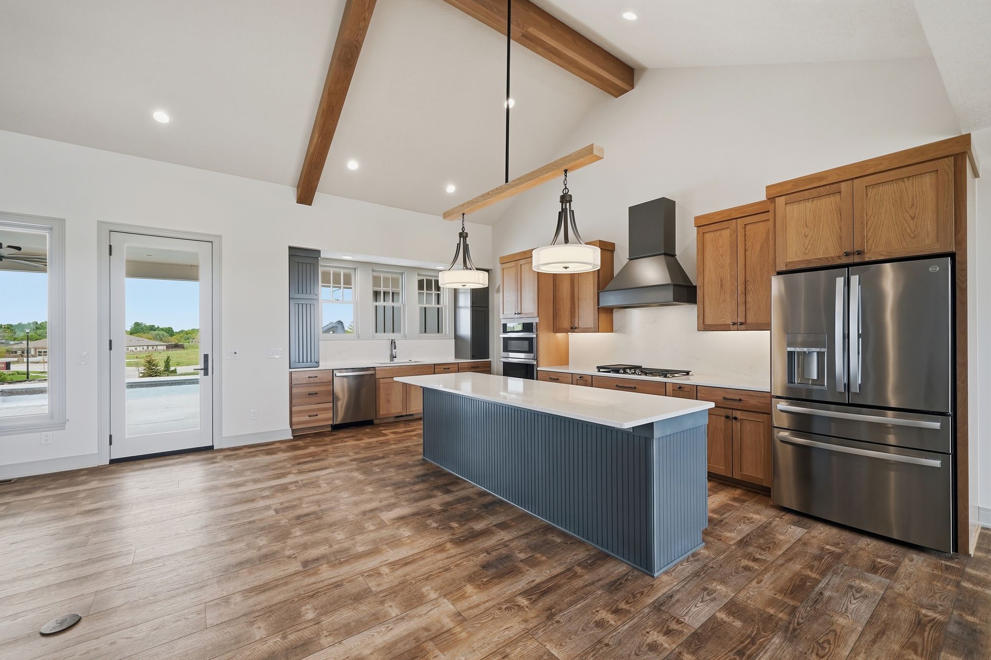Spacious kitchen with vaulted ceiling, wood beams, and wood-look flooring. A gray-blue kitchen island sits between stainless steel appliances, wood cabinets, and a double-door, glass door leading to the outside.