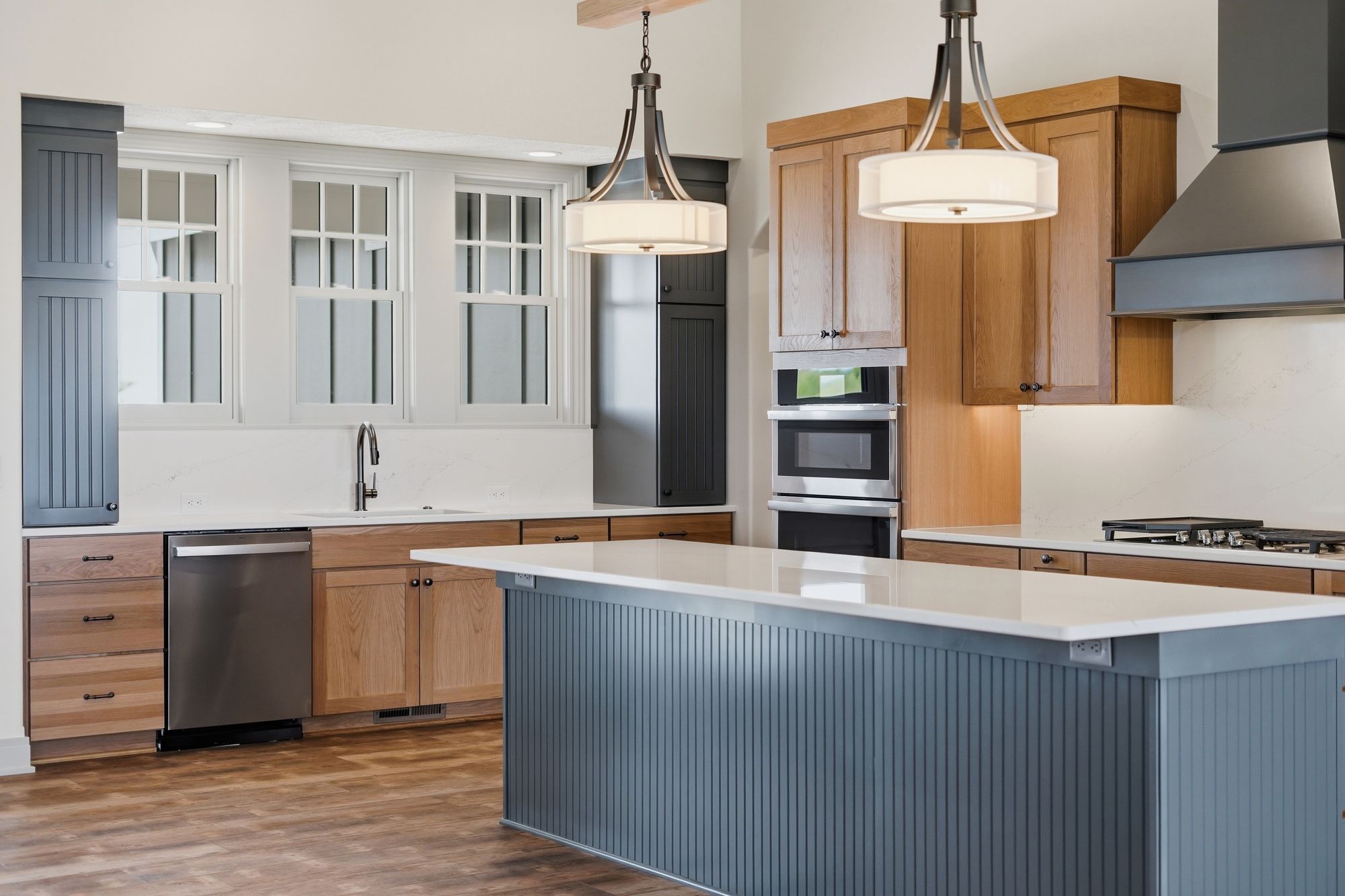 Modern kitchen featuring oak cabinetry, stainless steel appliances, and a blue gray island with white countertops. Pendant lighting hangs above the island, illuminating the space with natural light from nearby windows.