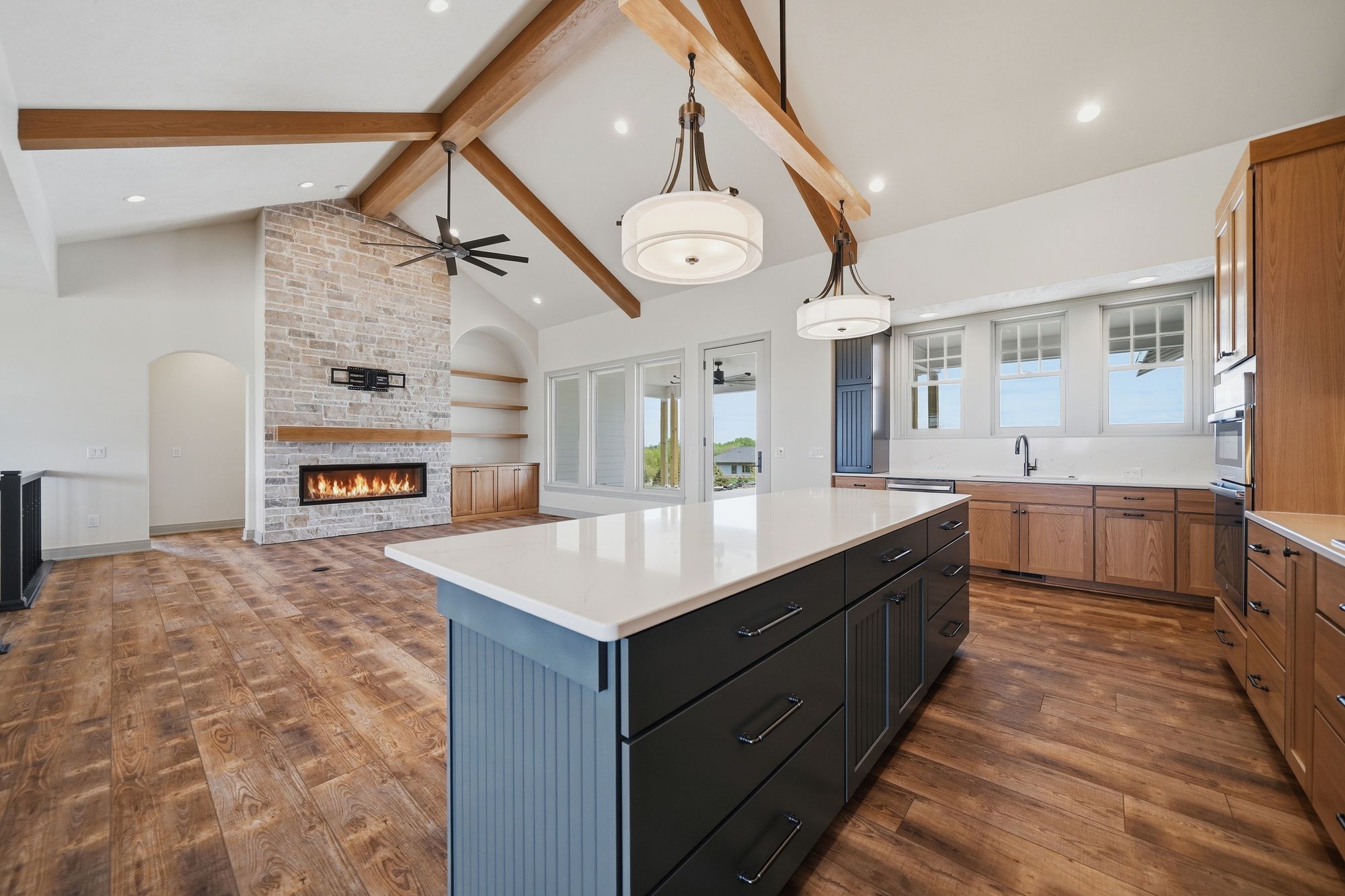 Open concept kitchen and living area featuring hardwood floors, a brick fireplace with a wood mantel, and an island with a white countertop. The kitchen has light wood cabinets with white countertops and a window above the sink.