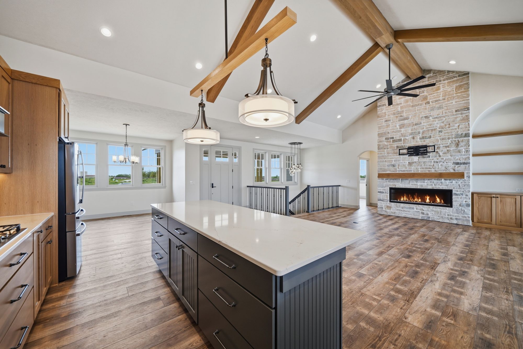 Open concept kitchen and great room showcase hardwood flooring and natural light. The kitchen features a large island with white countertops and dark cabinetry, while the great room has a stone fireplace and vaulted ceiling with exposed beams.
