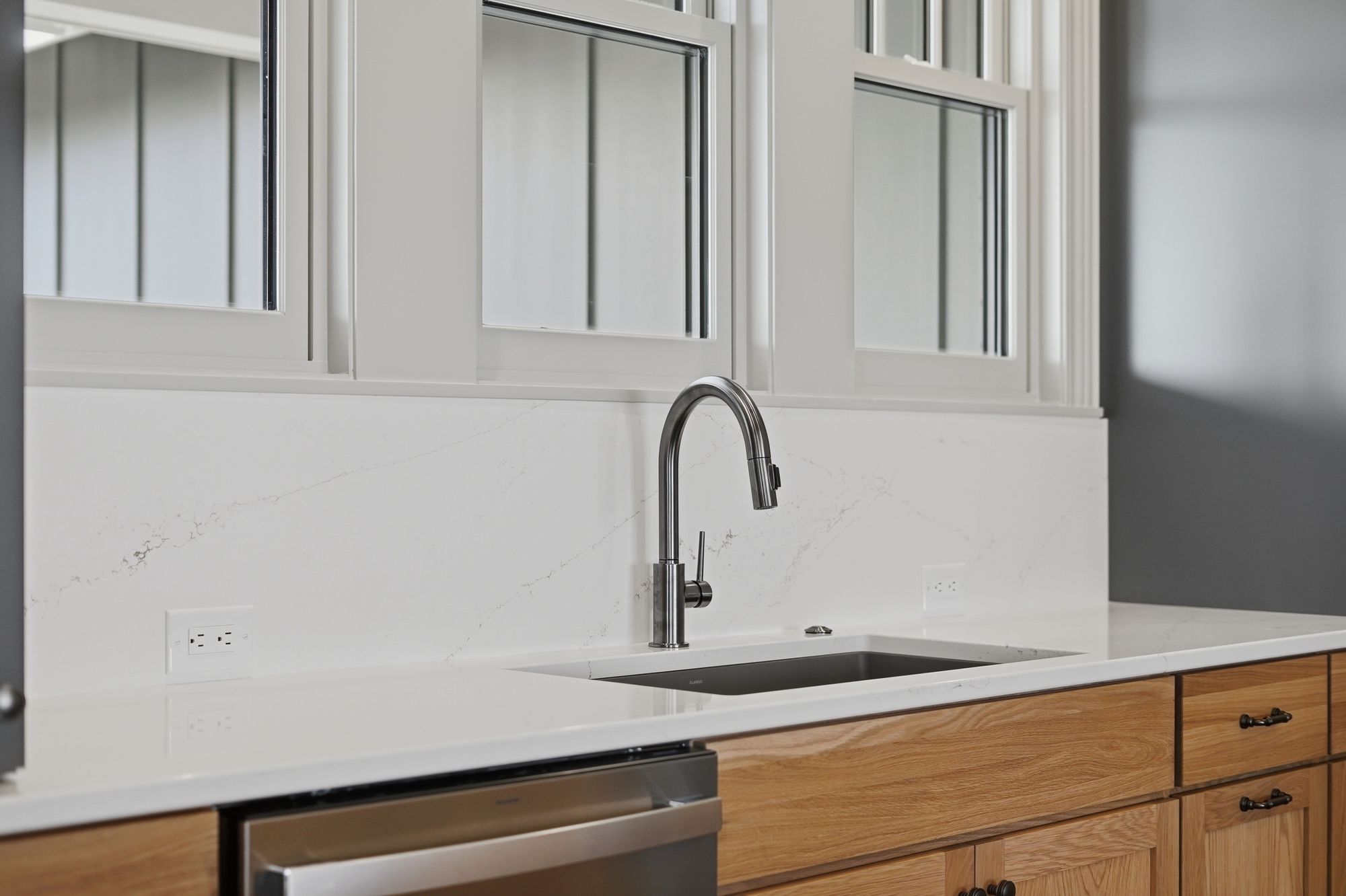 Modern kitchen features a stainless steel faucet and sink, light wood cabinets, and white countertops with subtle veining. Three windows provide natural light above the countertop.