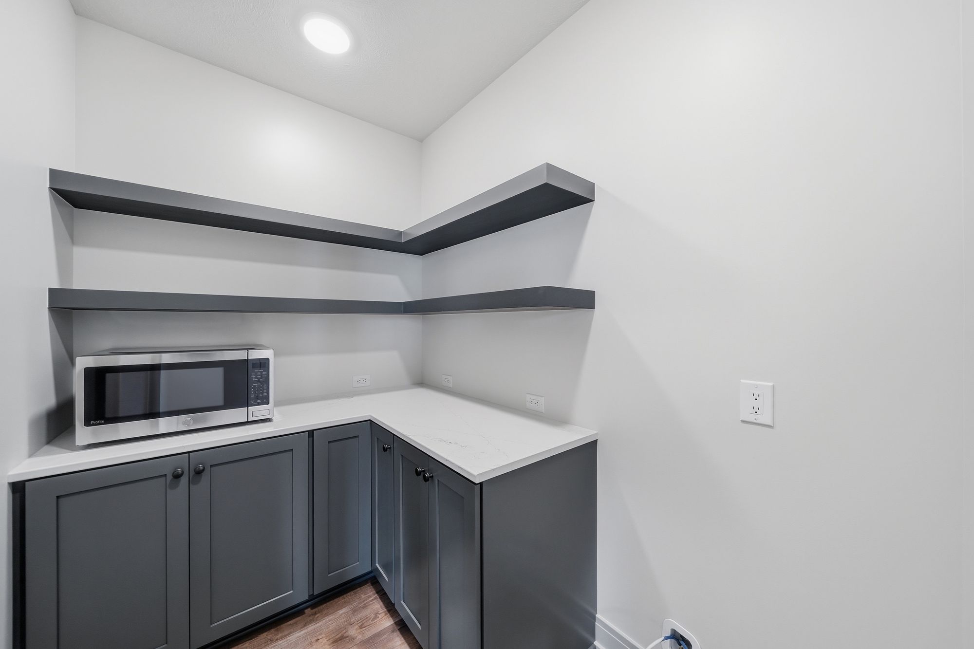 Pantry area featuring gray cabinets with a white countertop, and a microwave placed in the corner. Above the countertop are floating gray shelves creating corner storage.