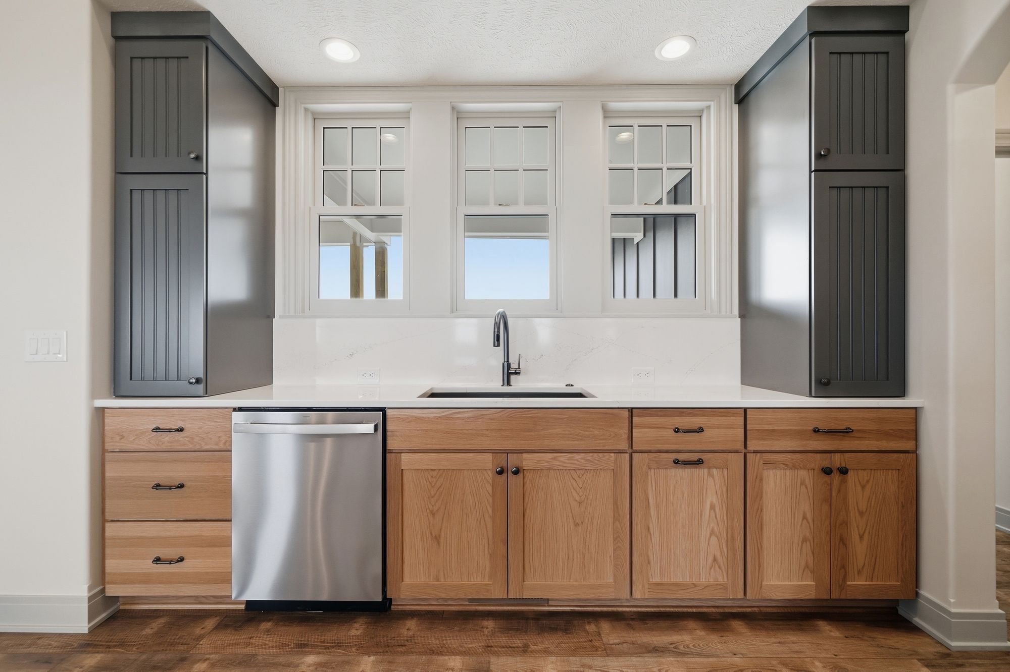 This wet bar features light wood cabinetry with dark bronze hardware, paired with a white quartz countertop and a stainless steel dishwasher. Three white-framed windows offer natural light above the counter.