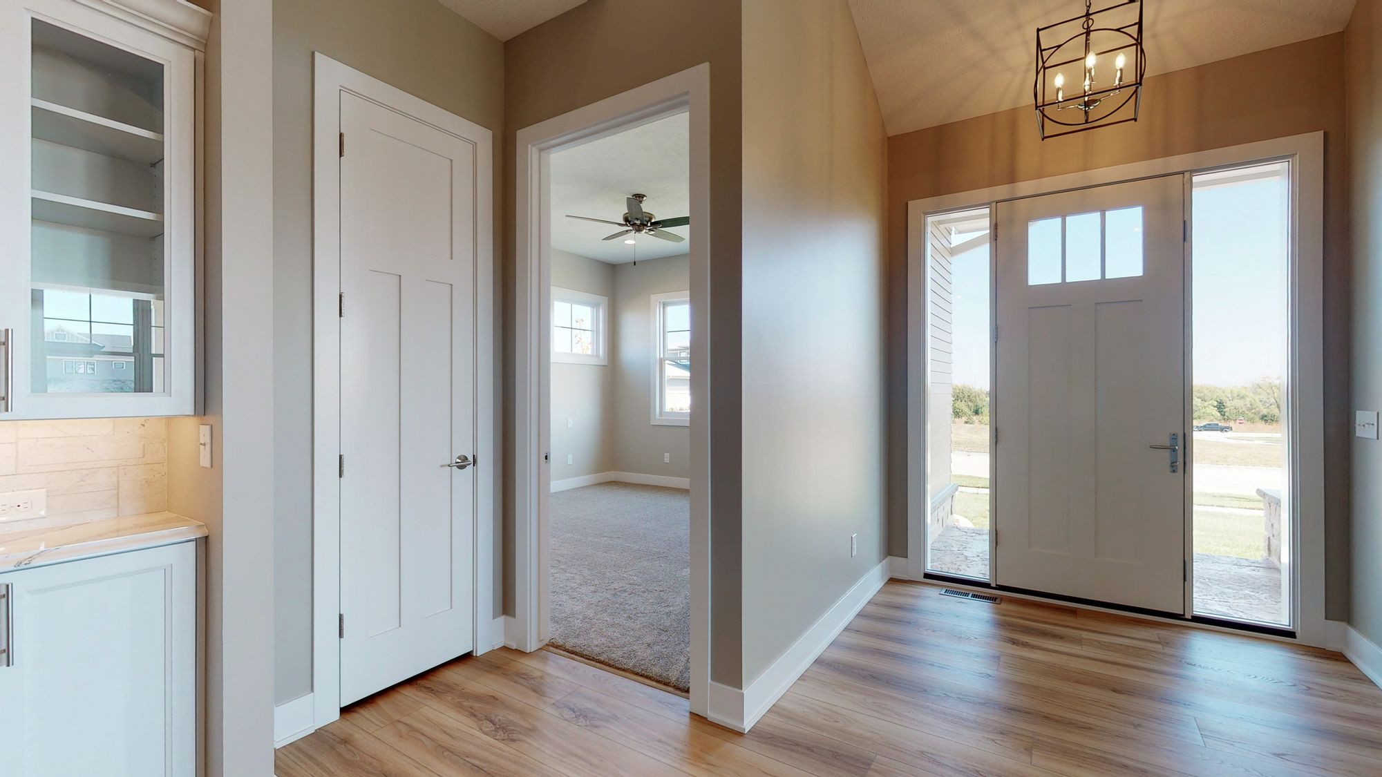 Entryway featuring light wood-look flooring, a white front door with sidelights, and an adjacent doorway leading to a carpeted room. A white painted door to the left offers storage space.