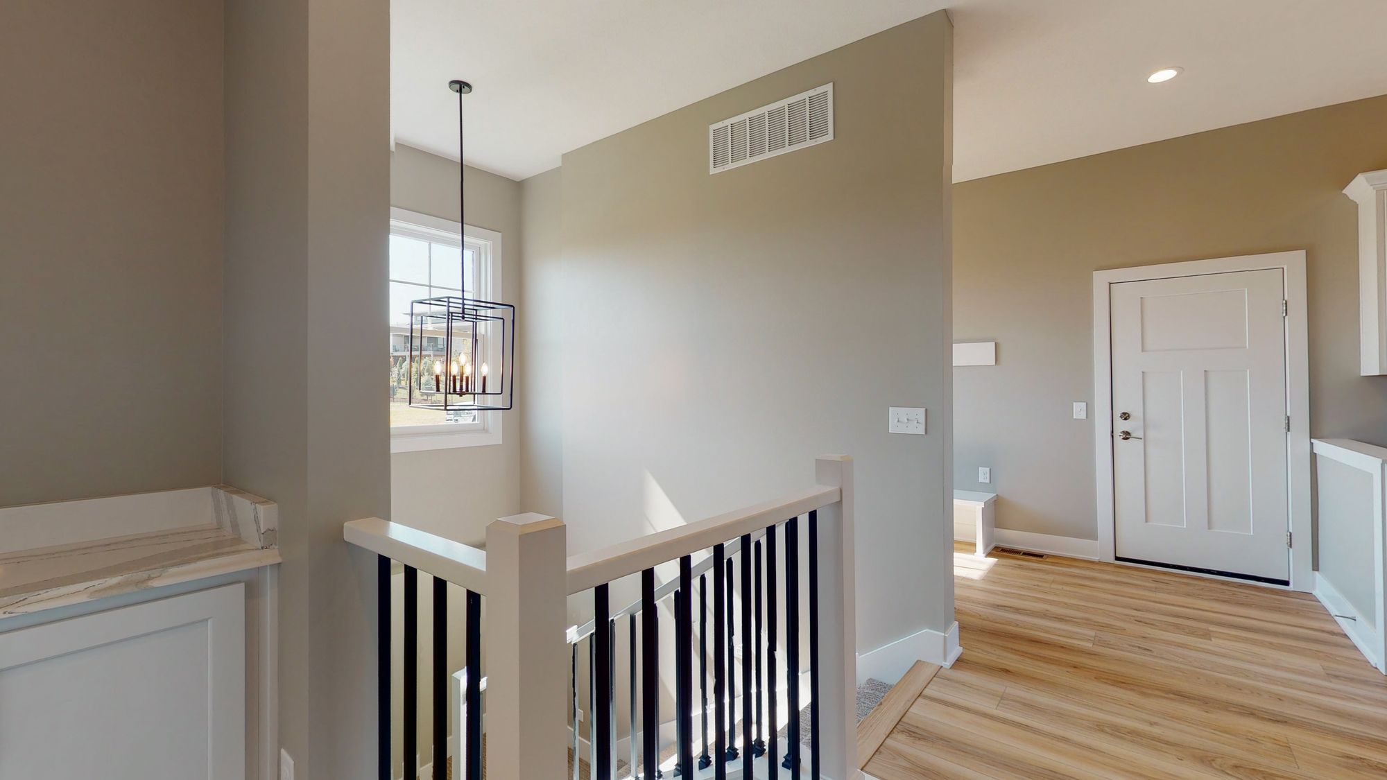 Interior view of a home's hallway featuring a staircase with white railings and black balusters, a modern chandelier, and a glimpse into a room with light wood floors and a white door. The walls are painted in a neutral tone, and natural light fills the space through a window.