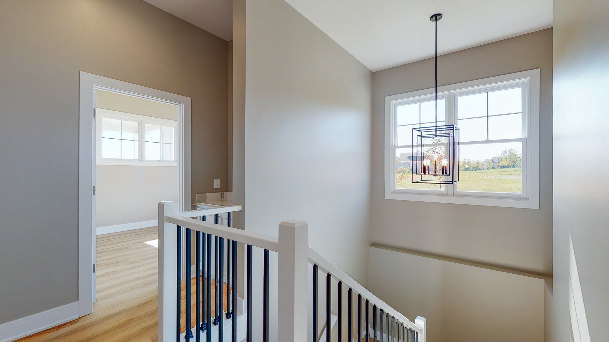 Interior shot of a stairway landing area featuring neutral-toned walls, white-and-black railing, and a modern geometric chandelier hanging in front of a window with a view of a green lawn and houses. An open doorway leads to another room with light-colored wood floors.