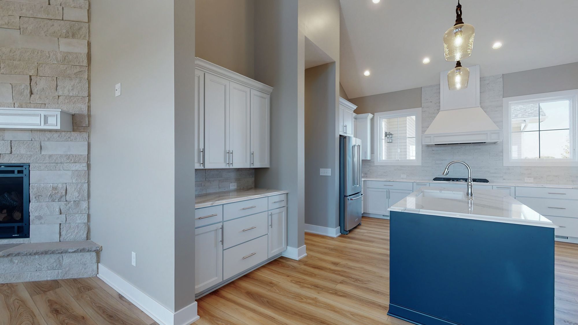 Bright kitchen featuring a dark blue island with marble countertops, white cabinetry, stainless steel appliances, and hardwood flooring. A stone fireplace with a white mantel provides a contrasting texture.