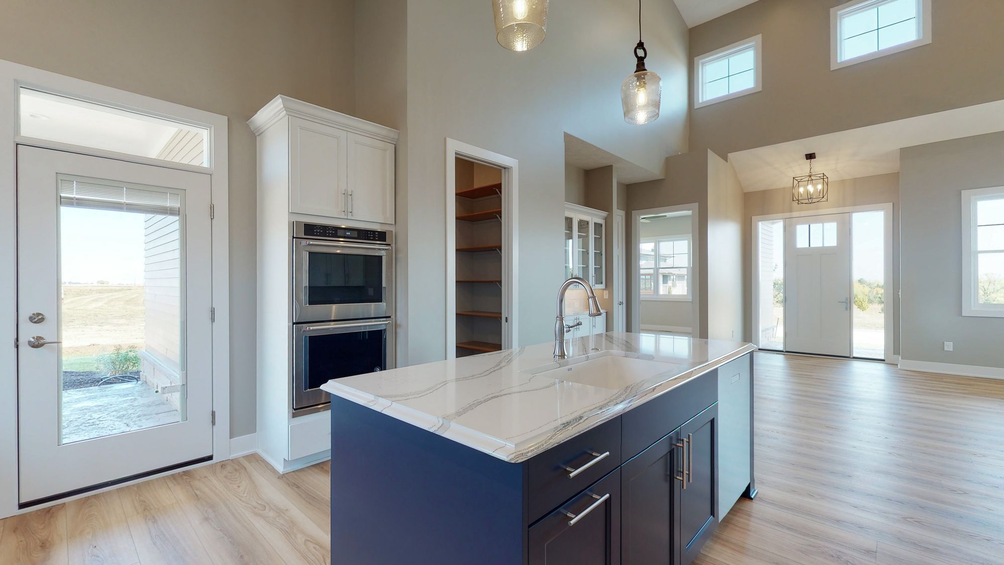 Bright and spacious kitchen featuring a blue island with white marble countertop and stainless steel appliances. The room is adjacent to a walk-in pantry and has natural light from windows and a door to the patio.
