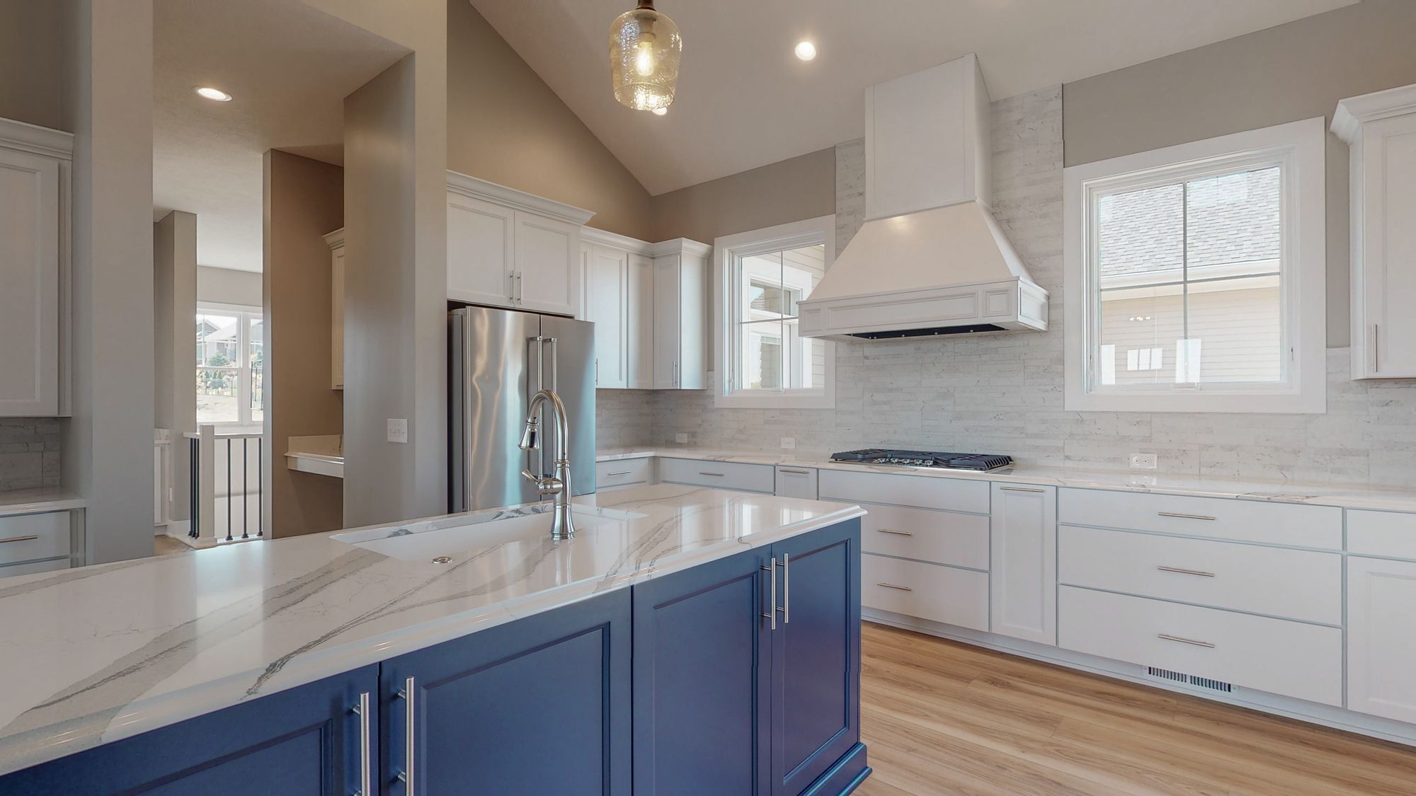 Modern kitchen featuring a large marble island with blue cabinets, white perimeter cabinets, and stainless steel appliances. The space has wood flooring and a light-colored backsplash.