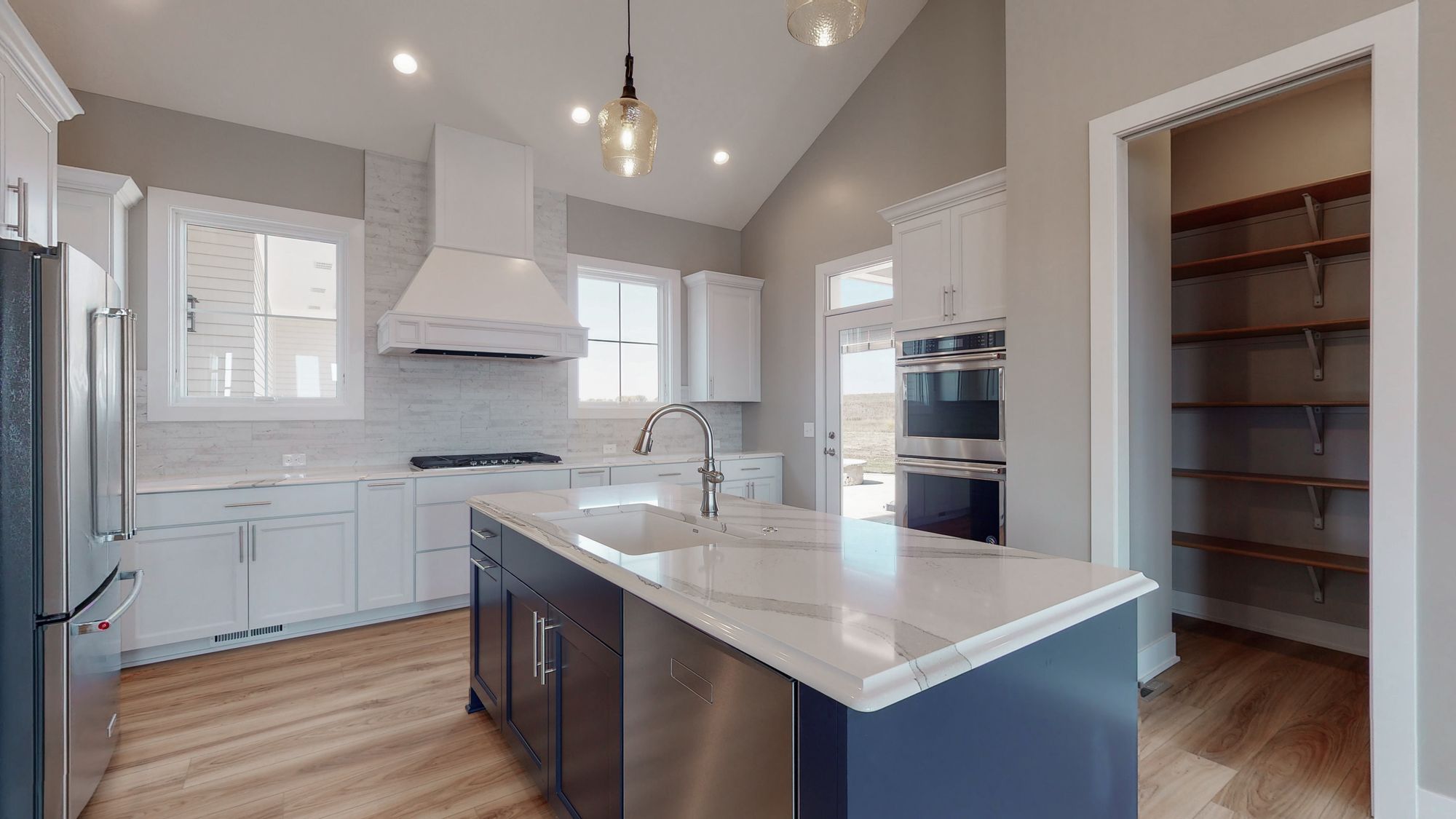 Bright kitchen with white cabinets and a navy blue island featuring a marble countertop. The kitchen includes stainless steel appliances and access to a pantry with wooden shelving.