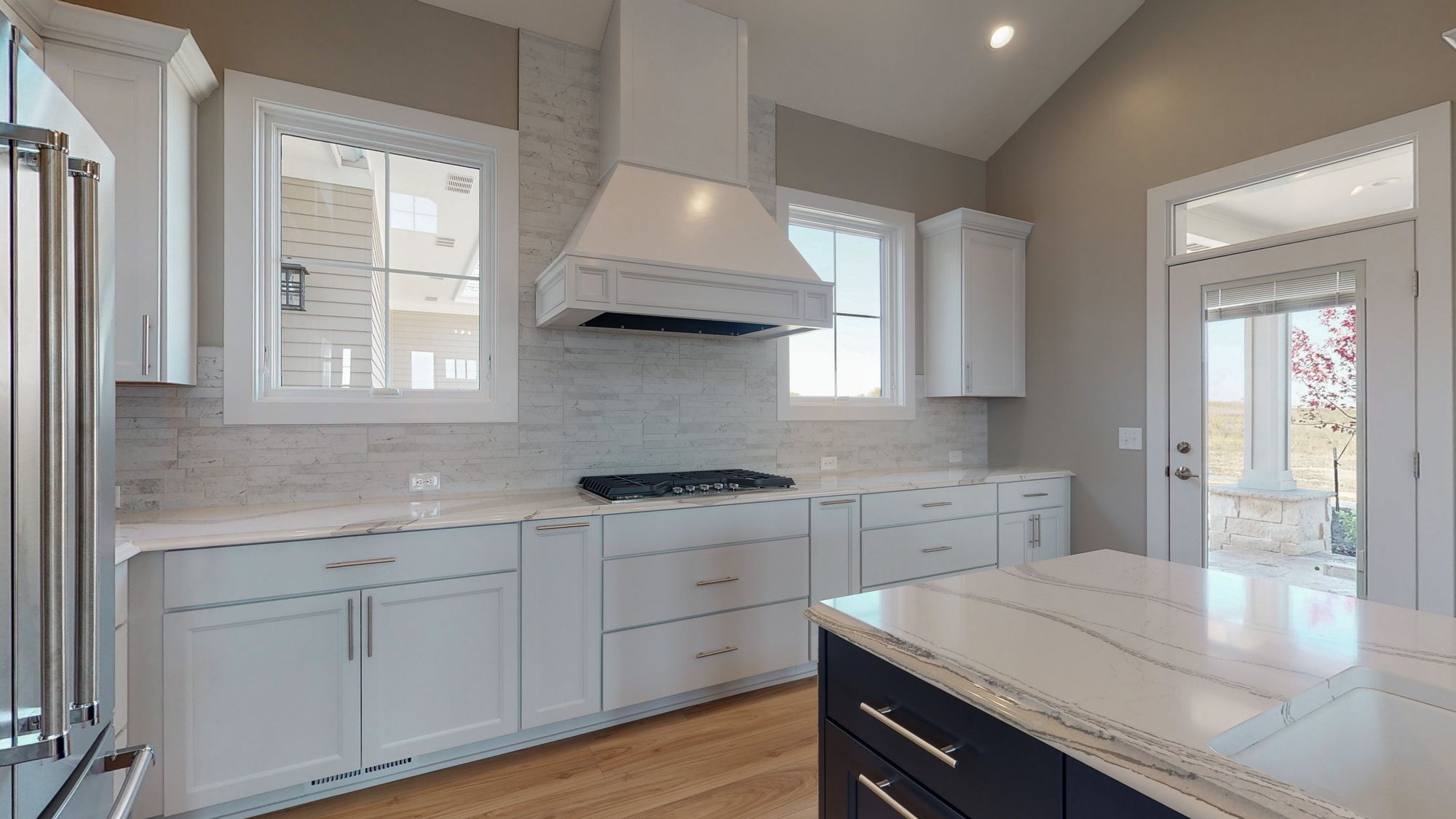 Modern kitchen featuring white shaker cabinets, a marble island with a contrasting dark base, and stainless steel appliances. A large window provides natural light, complementing the light wood flooring.