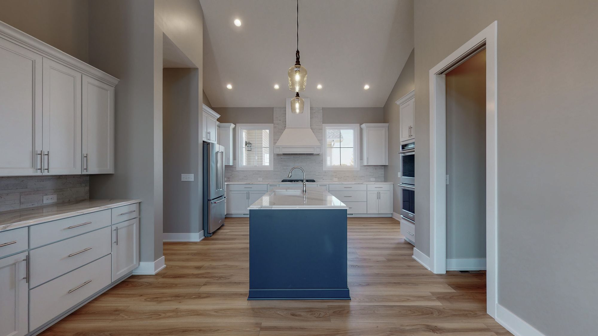 Modern kitchen features white cabinetry, a navy island with a marble countertop, and stainless steel appliances. The open-concept layout includes wood flooring and vaulted ceilings with pendant lighting.