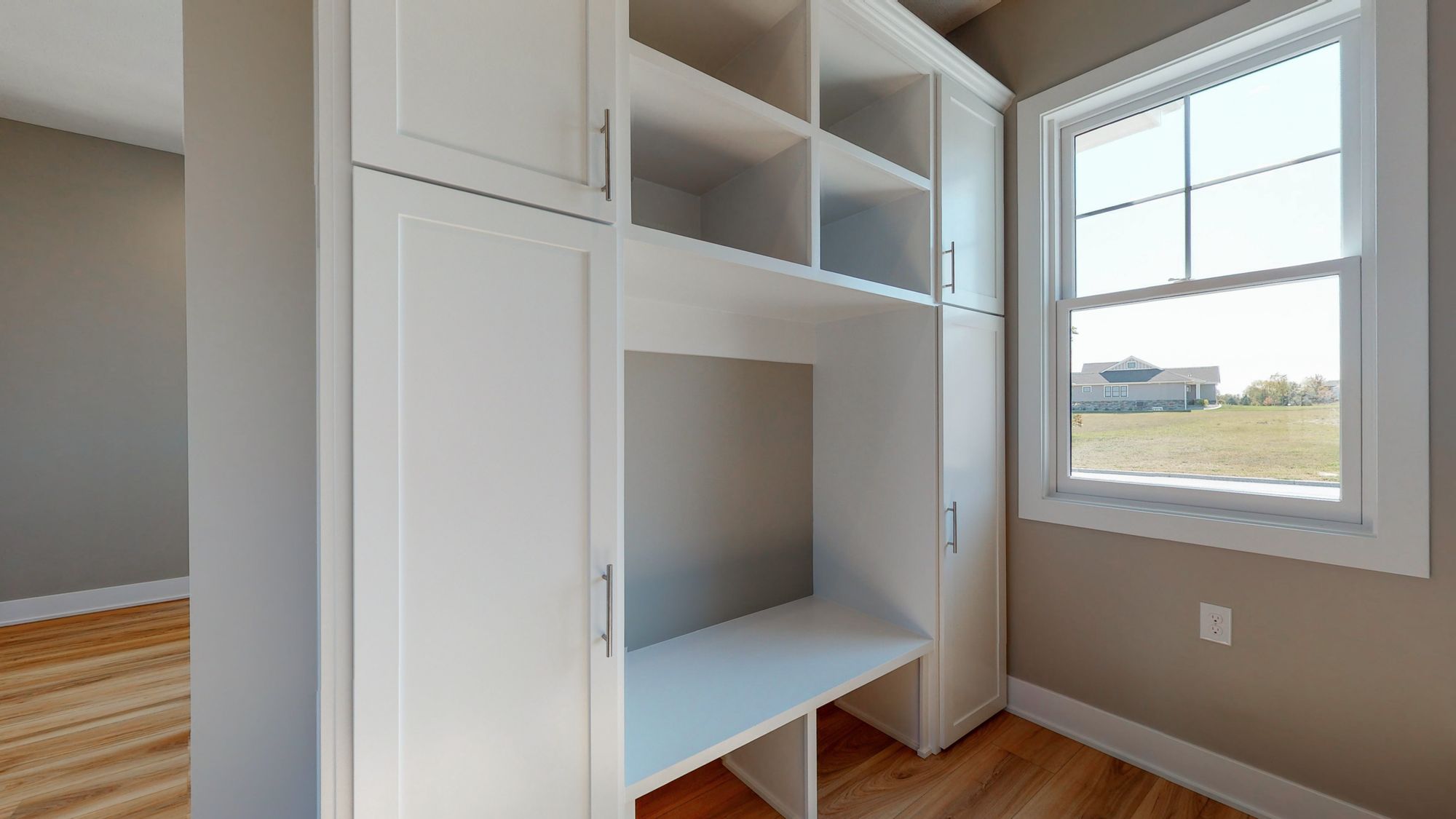This is a mudroom with built-in white cabinetry, including shelving and a bench. The room features hardwood floors and a window with views of a house and yard.
