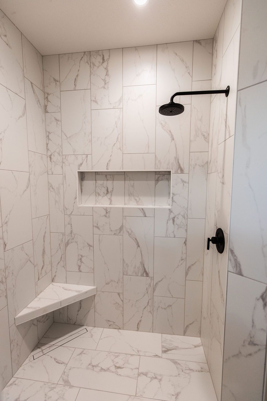 Marble-tiled shower with black hardware including a shower head and valve. The shower features a corner seat and a niche for storage.