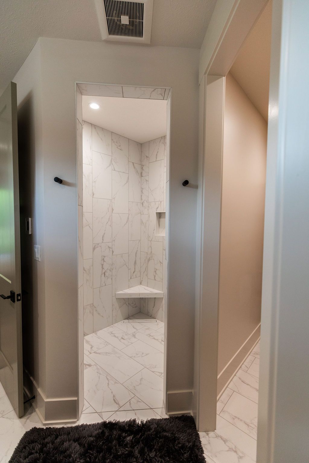 Modern bathroom featuring a walk-in shower with marble-patterned tile walls and flooring. A corner bench provides seating, complemented by matte black towel hooks and a doorway leading to another room with a consistent marble tile design.