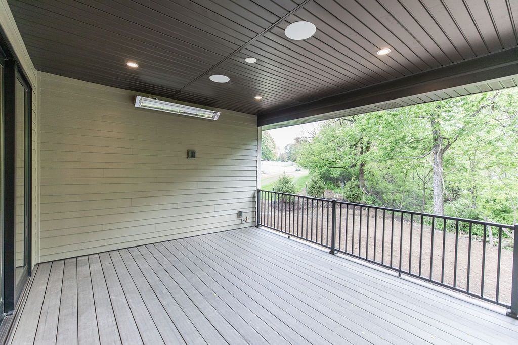 Covered deck area with grey flooring, horizontal siding, and a view of the yard with trees and a black metal railing. Overhead, a brown paneled ceiling features recessed lighting and a wall-mounted heater.