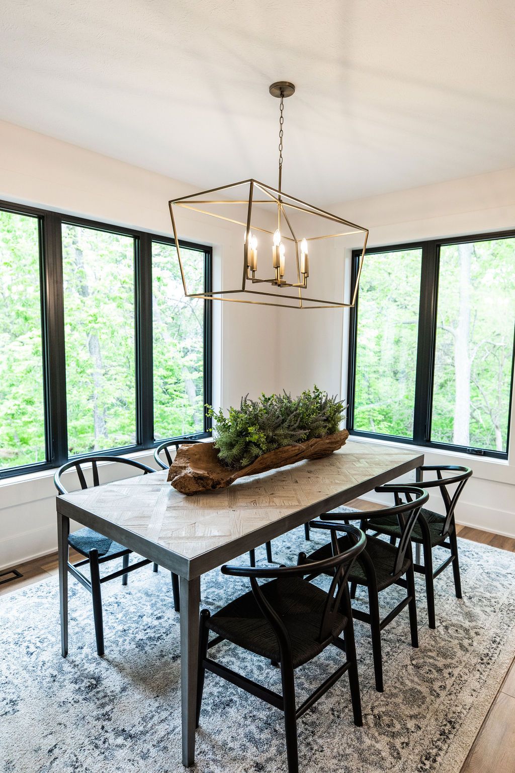 Modern dining room features a light wood dining table with a driftwood centerpiece, surrounded by black wishbone chairs on a gray patterned rug. A contemporary chandelier hangs above the table, with large windows offering views of the green trees.