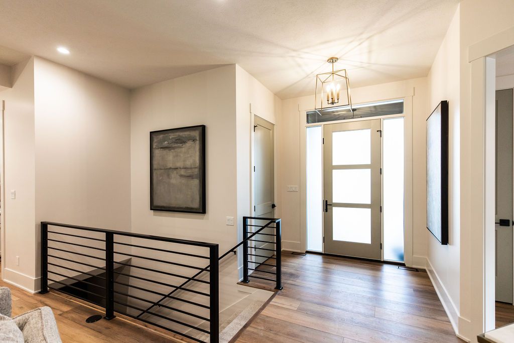 Entryway features a modern front door with frosted glass panels and sidelights, illuminated by a gold geometric chandelier. A black metal railing overlooks a lower level while a framed artwork hangs on the wall.