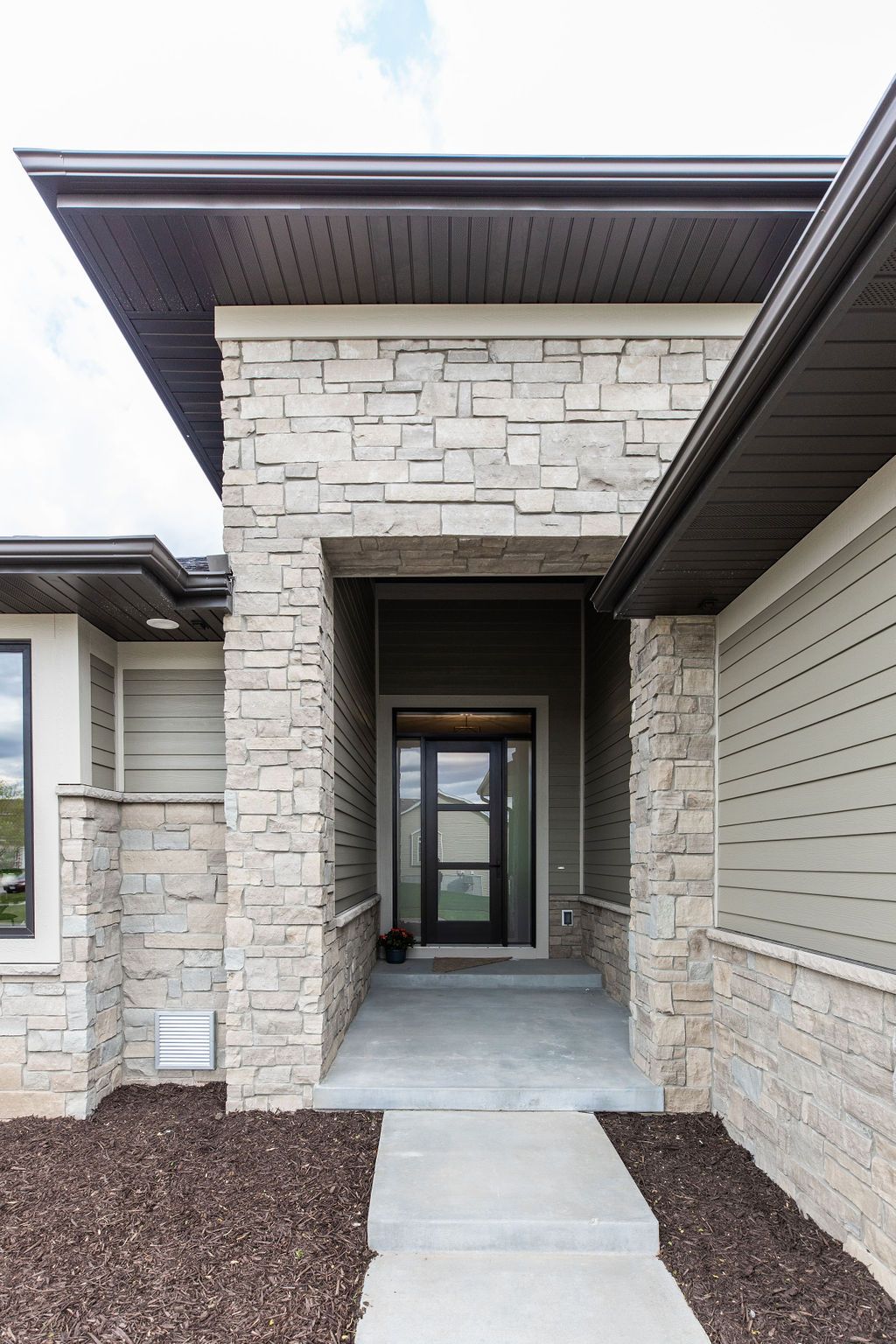 Exterior front entrance of a modern home featuring a stone veneer and horizontal siding. The entrance includes a concrete walkway, a dark-framed front door with a screen, and an overhanging roof.
