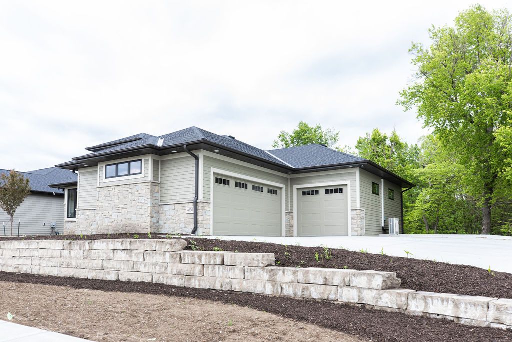 Exterior shot of a single-story home with a two-car garage and stone veneer accents. The house features light green siding and a dark gray roof, with a retaining wall visible in the front yard.