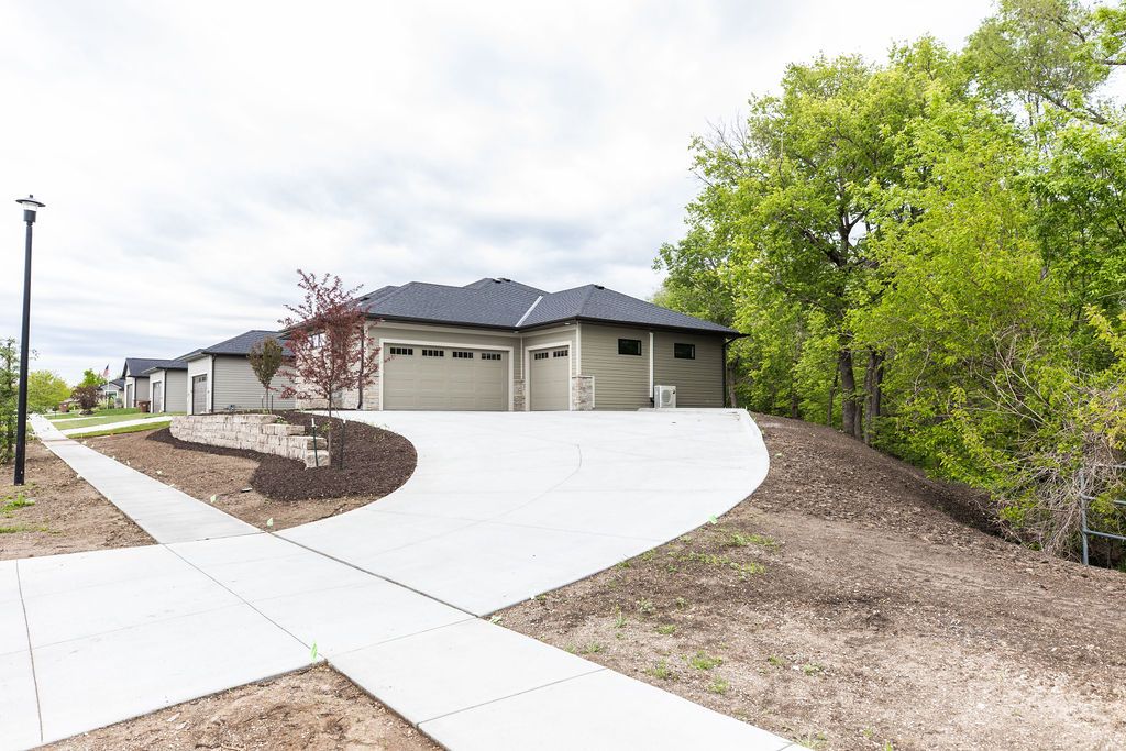 Exterior view of a modern, two-car garage house with a curving white driveway and a paved sidewalk. The house features light tan siding, a dark gray roof, and stone veneer accents around the garage doors.