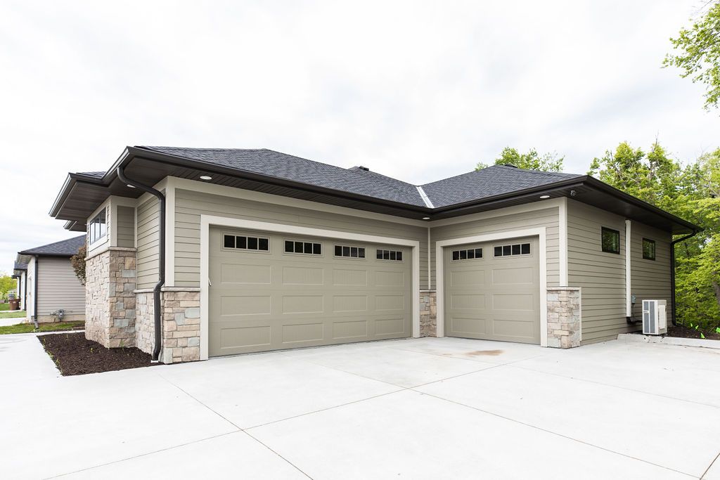 Exterior view of a modern home features a two-car garage, beige horizontal siding, and stone veneer accents. A concrete driveway leads to the garages, and a dark asphalt roof is visible overhead.