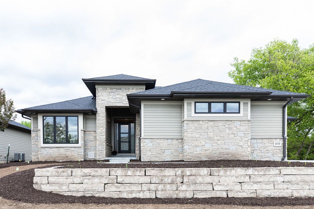 Modern single-story home with stone veneer accents and horizontal siding. Features include a dark gray gabled roof, black-framed windows, and a raised stone retaining wall landscaping.