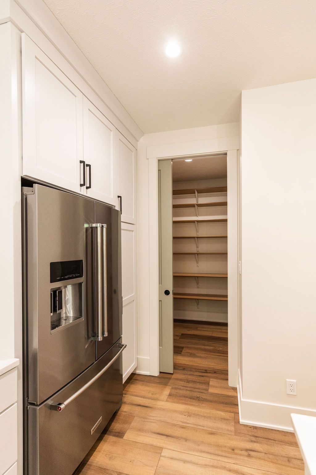 Kitchen area with stainless steel refrigerator adjacent to walk-in pantry with open shelving. Wood-look flooring complements the bright white walls and cabinetry.