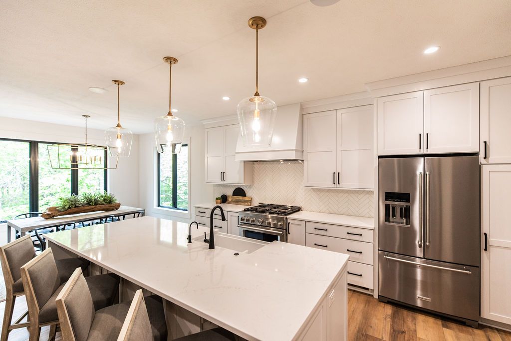 Bright and airy kitchen with white cabinetry, a large center island with a white countertop, and stainless steel appliances, complemented by modern pendant lighting and hardwood floors. A dining area with a long table and chairs sits adjacent to the kitchen, illuminated by large windows.