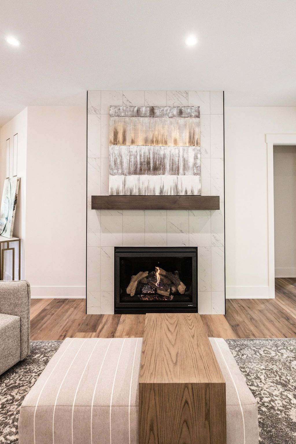 Living room featuring a fireplace with marble-look tile surround and a dark wood mantel, topped by an abstract artwork. A striped ottoman and wooden coffee table sit on a patterned rug in front of the fireplace.