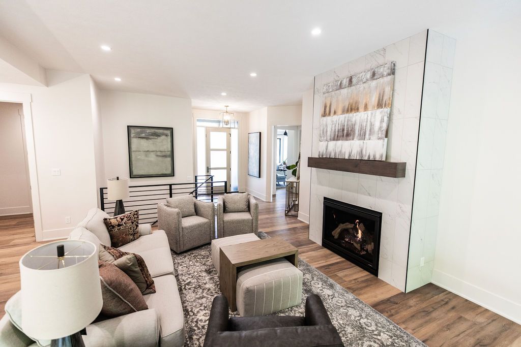 Bright living room showcasing a modern fireplace with marble tile and a dark wood mantle, flanked by a light-colored sofa and two armchairs. An area rug anchors the space with a neutral color palette, and recessed lighting illuminates the room.