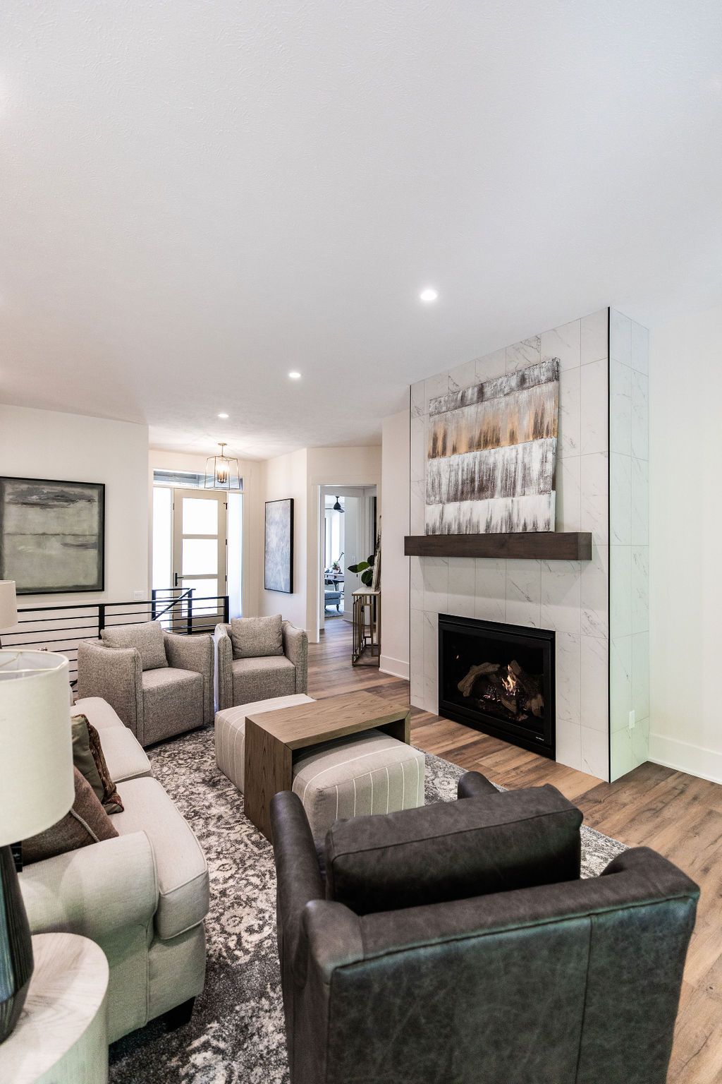 Bright living room features a fireplace with light marble surround, mantel, and modern artwork above. Furniture includes a light-colored sofa, two gray armchairs, and a dark gray armchair, centered around a patterned rug and coffee table.