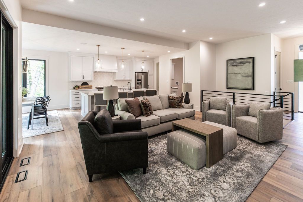 Open-concept living room and kitchen feature hardwood floors, recessed lighting, and a gray color palette. The living area includes a gray sofa, two armchairs, an ottoman, and a patterned rug, while the kitchen has white cabinets, stainless steel appliances, and a central island with bar seating.