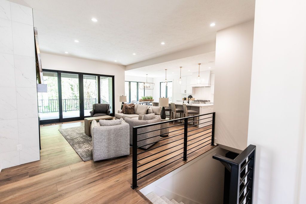 Open concept living room and kitchen area featuring light gray upholstered sofas, wood floors, and a black metal railing along the staircase. The adjacent kitchen has white cabinetry and a large center island.
