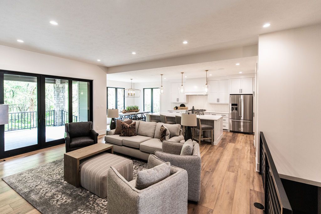 Open-concept living space features hardwood floors, gray area rug, and comfortable seating. The kitchen with white cabinets and stainless steel appliances is visible beyond the living room.