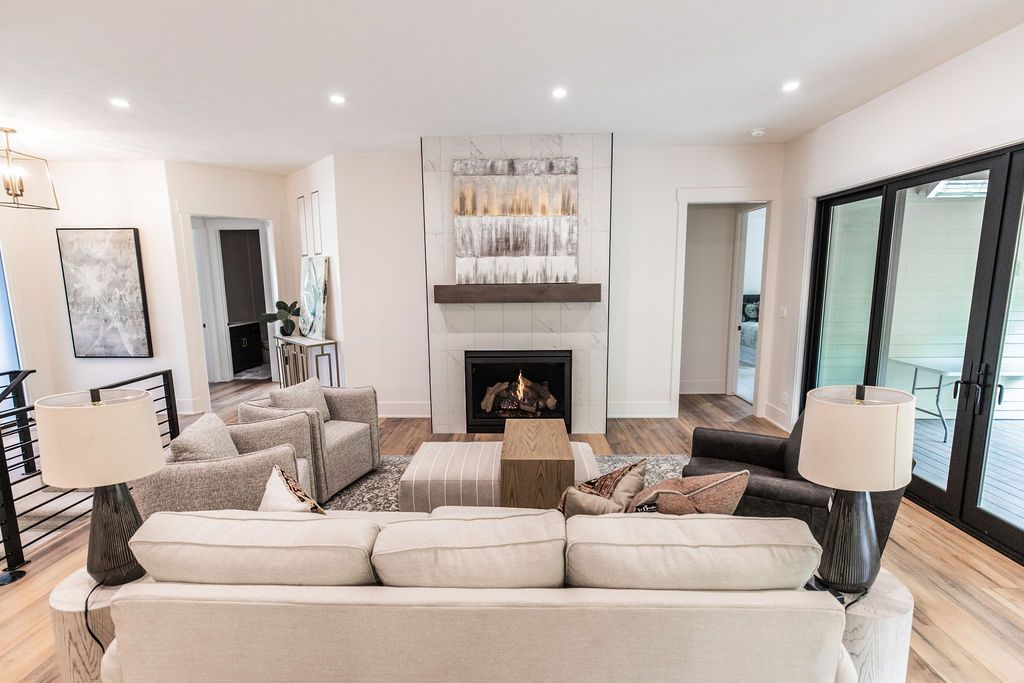 Modern living room with a fireplace, light gray sofa, and two accent chairs in light gray and dark gray. The room features a modern fireplace with a marble surround and a painting above the mantel, wood flooring, and black-framed sliding glass doors leading to an outdoor deck.