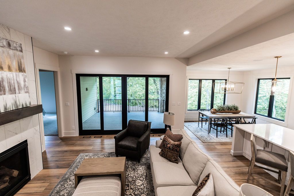 Interior view of an open-concept living and dining area, featuring hardwood flooring, a fireplace with a marble surround, a white sectional sofa, and a black-framed sliding glass door leading to a deck. The dining area has a table with a wooden centerpiece and modern black chairs.